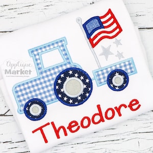 May include: A blue and white gingham tractor with a red, white, and blue American flag on the back. The tractor has a white star on each tire. The name Theodore is embroidered in red below the tractor.