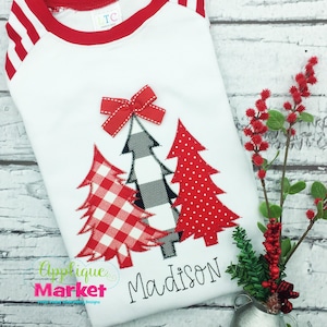 May include: White long-sleeve shirt with red and white striped sleeves and a red ribbed neckline. The shirt features an applique of three Christmas trees with a red bow on top. The trees are decorated with red and white gingham, black and white buffalo plaid, and red polka dots. The name "Madison" is embroidered below the trees.