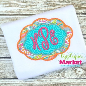 May include: A white t-shirt with a turquoise, pink, and orange embroidered monogram in a scalloped frame. The frame has a paisley pattern. The text "Applique Market" is below the monogram.