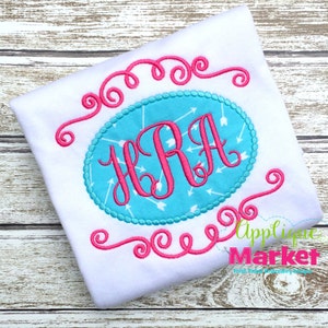 May include: A white t-shirt with a pink monogram in an oval frame with a turquoise background and white arrows. The frame is decorated with pink swirls.