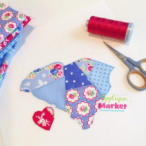 May include: A patchwork of blue and pink fabric in the shape of a heart, with a smaller red heart with a white deer design in the center. There is a spool of red thread, a pair of scissors, and a stack of fabric scraps in the background. The text "Applique Market Fresh Picked Embroidery Designs" is visible in the bottom right corner.