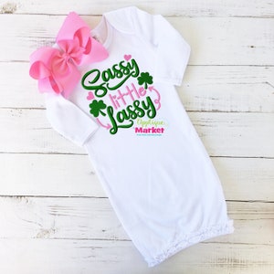 May include: White baby gown with a green and pink embroidered design that says "Sassy little Lass" with shamrocks and hearts.