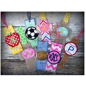 May include: A collection of nine colorful fabric bookmarks with various designs, including a cross, a baseball bat, a soccer ball, a football, and an apple. Each bookmark features a personalized monogram.
