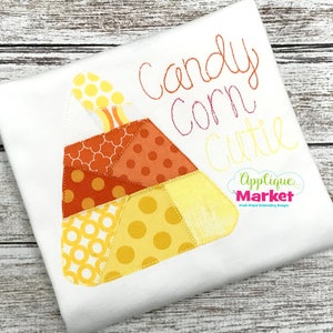 Puede incluir: Una camiseta blanca con una aplicación de un caramelo de maíz hecho de recortes de tela en amarillo, naranja y marrón. El texto "Candy Corn Cutie" está bordado sobre la aplicación. El texto "Applique Market" está bordado en la esquina inferior derecha.