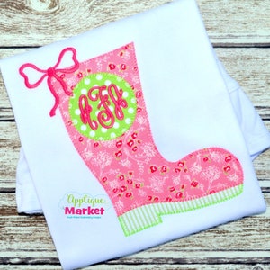 Può includere: Un'applique in tessuto rosa e verde a forma di stivale da pioggia con un fiocco e un monogramma in un cerchio. L'applique è su una maglietta bianca. Il testo "Applique Market" è nell'angolo in basso a sinistra dell'immagine.