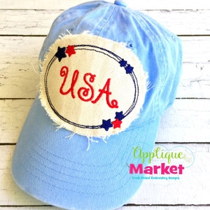 Puede incluir: Una gorra de béisbol azul claro con un parche de tela blanca que presenta un "USA" bordado en rojo con un diseño de estrellas azules y rojas.