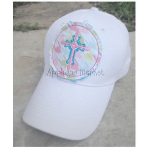 May include: White baseball cap with a round, colorful patch featuring a pink cross with blue accents. The patch has a distressed, frayed edge.
