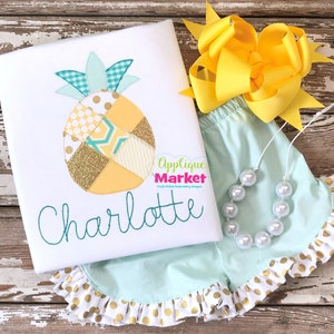 May include: A white t-shirt with a pineapple applique in yellow, gold, and teal, with the name "Charlotte" embroidered in teal. The outfit includes mint green shorts with gold polka dot ruffles, a yellow bow, and a pearl necklace.