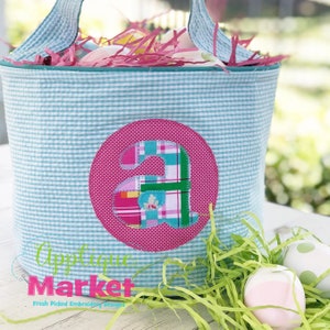 May include: A light blue Easter basket with a pink circle applique featuring the letter "A" in a patchwork design. The basket is filled with pink and white shredded paper and is sitting on a wooden table with green shredded paper and pink and white Easter eggs.