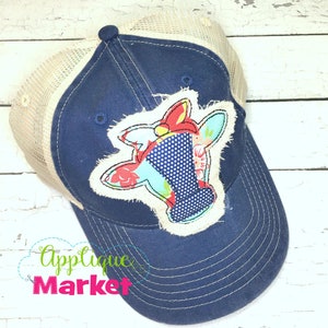 May include: A blue and beige trucker hat with a colorful cow applique. The applique is made of fabric scraps and has a red bow. The hat has a mesh back and a curved brim.