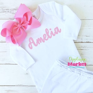 Op de afbeelding: Witte romper met lange mouwen met de naam "Amelia" geborduurd in roze op de voorkant. Een roze strik zit op de linker schouder.