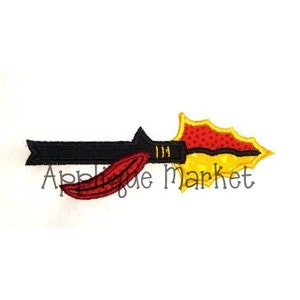 Puede incluir: Una aplicación bordada de una lanza con un eje negro, una pluma roja y una punta amarilla. El número "111" está bordado en el eje.