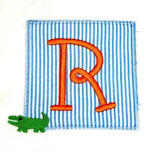 Op de afbeelding: Een blauw en wit gestreept vierkant van stof met een grote, oranje geborduurde letter "R" en een groene alligator-applicatie.