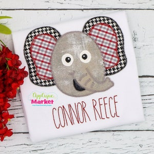 Puede incluir: Camiseta blanca con una aplicación de elefante gris con orejas a cuadros rojos y blancos. El elefante tiene ojos negros y una nariz cosida negra. El texto "CONNOR REECE" está bordado en rojo debajo del elefante.