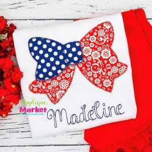 Può includere: Una maglietta bianca con un'applicazione a fiocco rosso e blu e il nome "Madeline" ricamato in filo blu.