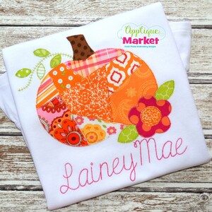 May include: A white t-shirt with a patchwork pumpkin applique in shades of orange, pink, and green. The pumpkin has a brown stem and a green vine with leaves. A pink flower with orange and pink accents is next to the pumpkin. The name "Lainey Mae" is embroidered in pink below the pumpkin.