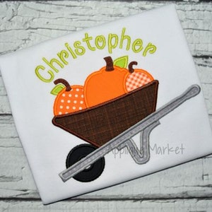 May include: A white t-shirt with the name "Christopher" embroidered in green. The shirt features an embroidered design of a wheelbarrow filled with orange, white, and red pumpkins.