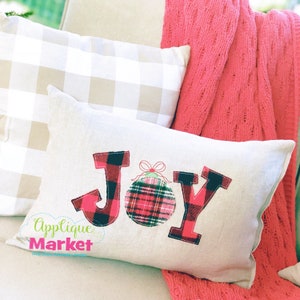 May include: A white pillow with the word "JOY" in red and black plaid fabric. The "O" is replaced with a red and black plaid ornament with a green bow. The pillow is on a white and brown gingham pillow and a pink knitted blanket.
