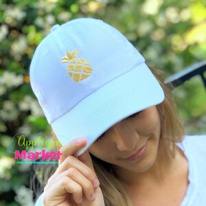 May include: A white baseball cap with a gold embroidered pineapple design.