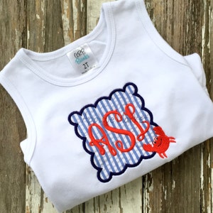 May include: White sleeveless shirt with a blue and white gingham monogram and a red crab appliqué. The monogram is in a navy blue scalloped frame. The shirt has a tag that says "Gap Blanks 2T Made in China".