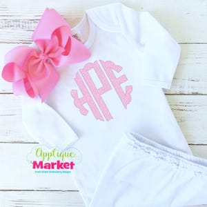 May include: A white baby gown with a pink monogram and matching pants. A large pink bow is placed next to the gown. The Applique Market logo is visible in the lower left corner.