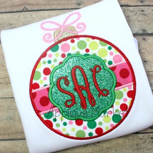 Op de afbeelding: Een rode, groene en roze kerstbal met een witte achtergrond. De kerstbal heeft een groen glittermonogram "SAC" in het midden.