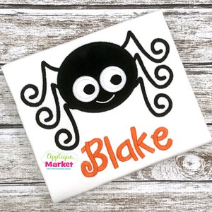 Puede incluir: Una camiseta blanca con una aplicación de araña negra y el nombre "Blake" en letras naranjas. La araña tiene grandes ojos blancos y una boca sonriente.