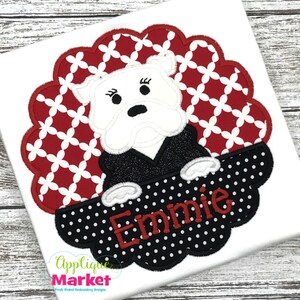 May include: Embroidered design featuring a white bulldog wearing a black jacket, peeking over a black and white polka dot banner with the name "Emmy" in red lettering. The background is a red and white geometric pattern.