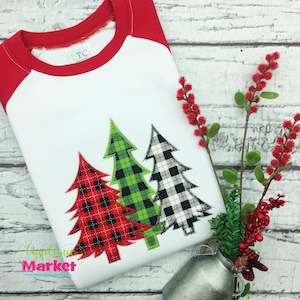 May include: A white, red, and green plaid Christmas tree applique on a white t-shirt with red sleeves. The t-shirt is on a white wooden background.