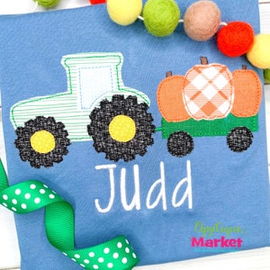 May include: A blue t-shirt with a green tractor pulling a wagon full of orange pumpkins. The tractor has black wheels and the wagon has two black wheels. The name "Judd" is embroidered in white on the shirt.