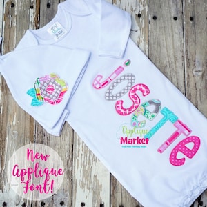 May include: A white baby gown with long sleeves and a matching hat. The gown has the name "Isabelle" embroidered on the front in a colorful applique font. The hat has a flower applique on the front.