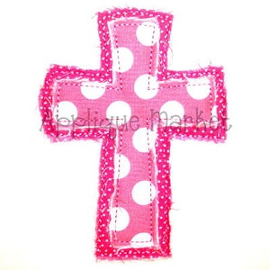 May include: A pink fabric cross with white polka dots. The cross has a frayed edge and is sewn with a pink thread.
