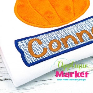 Puede incluir: Una camiseta blanca con una aplicación de calabaza naranja y una pancarta azul con el nombre "Connor" bordado en hilo marrón.