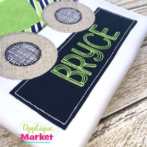 Puede incluir: Una camiseta blanca con un diseño de camión de aplique verde, azul y blanco con el nombre "BRYCE" bordado en verde sobre un rectángulo azul marino.