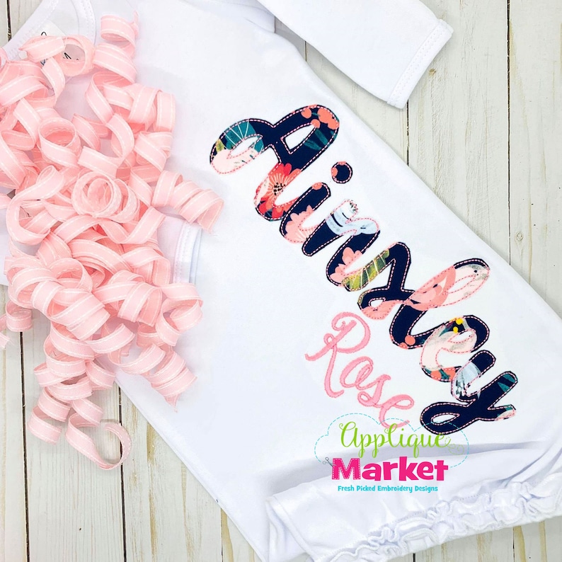 K&ouml;nnte beinhalten: Wei&szlig;er Baby-Schlafsack mit dem Namen "Ainsley Rose" in Rosa und Blumenstoff gestickt. Eine rosa Schleifenlocke befindet sich auf der linken Seite. Der Schlafsack hat lange &Auml;rmel und einen R&uuml;schensaum. Das Applique Market-Logo befindet sich unten.