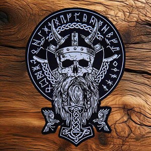 May include: A black and white Viking skull patch with a horned helmet, long beard, and axes. The design includes runic text around the edge and a hammer symbol at the bottom. The patch is circular and set against a wood grain background.
