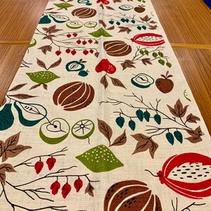May include: A white linen table runner with a repeating pattern of brown, green, and red fruits and leaves. The pattern includes pears, apples, pomegranates, and lemons.