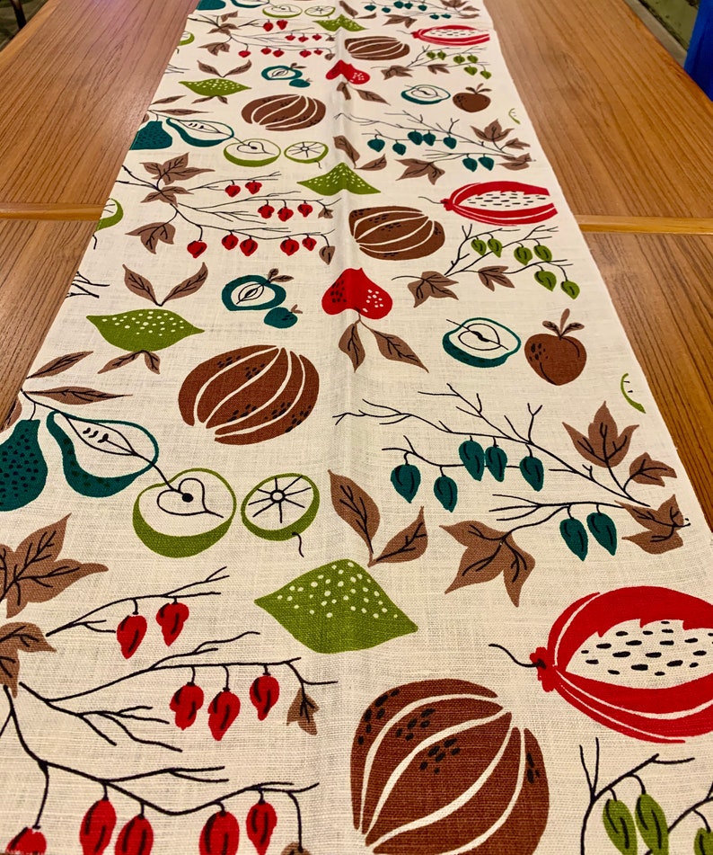 May include: A white linen table runner with a repeating pattern of brown, green, and red fruits and leaves. The pattern includes pears, apples, pomegranates, and lemons.