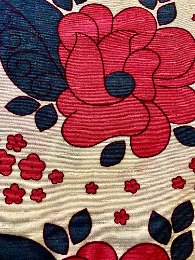 May include: A cream-colored fabric with a repeating pattern of red and blue flowers. The flowers are stylized and have a retro look. The fabric has a textured surface.