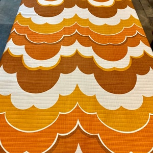 Op de afbeelding: Een retro stof met een herhalend wolkenpatroon. Het ontwerp toont lagen van oranje, bruine en gele tinten met witte contouren. De stof heeft een getextureerde uitstraling, wat wijst op een geweven materiaal, en is geschikt voor bekleding of woondecoratie.