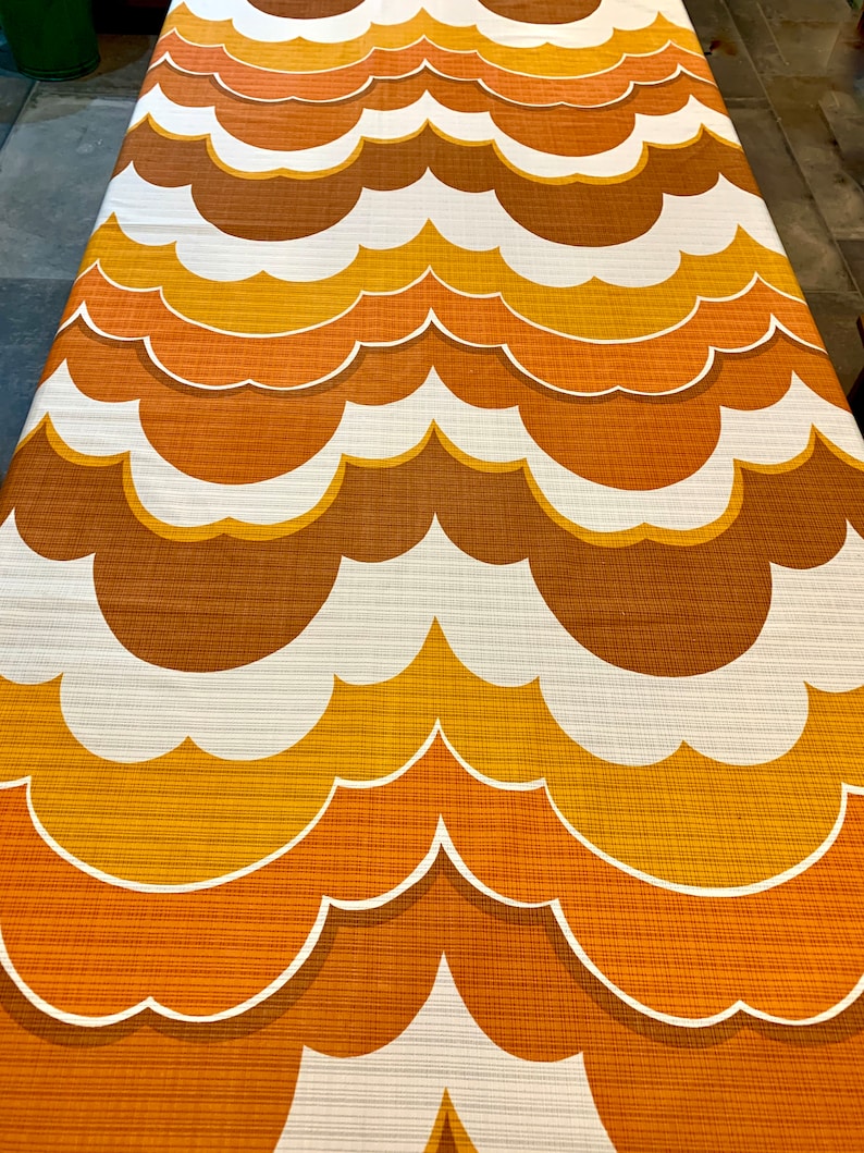 Op de afbeelding: Een retro stof met een herhalend wolkenpatroon. Het ontwerp toont lagen van oranje, bruine en gele tinten met witte contouren. De stof heeft een getextureerde uitstraling, wat wijst op een geweven materiaal, en is geschikt voor bekleding of woondecoratie.