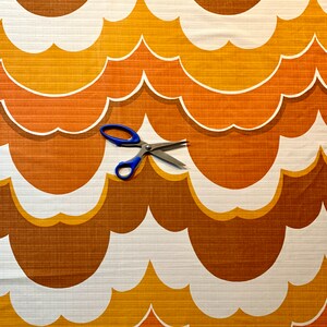 Op de afbeelding: Een retro-stof met een herhalend patroon van gestileerde wolkvormen in bruin, oranje en wit. Een blauwe schaar ligt op de stof. De stof heeft een getextureerd oppervlak en de afbeelding roept een retro-esthetiek op.