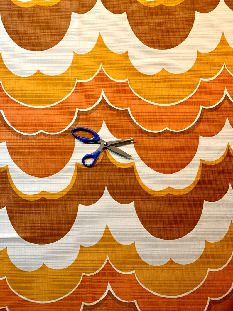 Op de afbeelding: Een retro-stof met een herhalend patroon van gestileerde wolkvormen in bruin, oranje en wit. Een blauwe schaar ligt op de stof. De stof heeft een getextureerd oppervlak en de afbeelding roept een retro-esthetiek op.