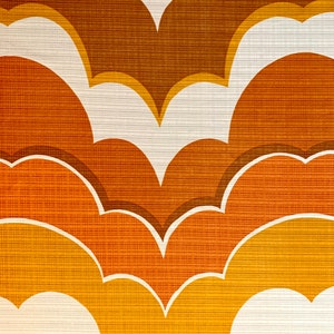 Op de afbeelding: Close-up van een retro behangpatroon. Het ontwerp toont gelaagde, wolkvormige vormen in tinten bruin, oranje en geel, omlijnd met wit. Het gestructureerde oppervlak voegt diepte toe aan het herhalende motief.