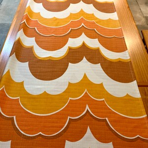 Op de afbeelding: Een tafelloper met een herhalend patroon van wolkvormige vormen in bruin-, oranje- en wittinten. De loper is gemaakt van een textuurstof en wordt gepresenteerd op een houten tafel. Het ontwerp roept een vintage esthetiek op.