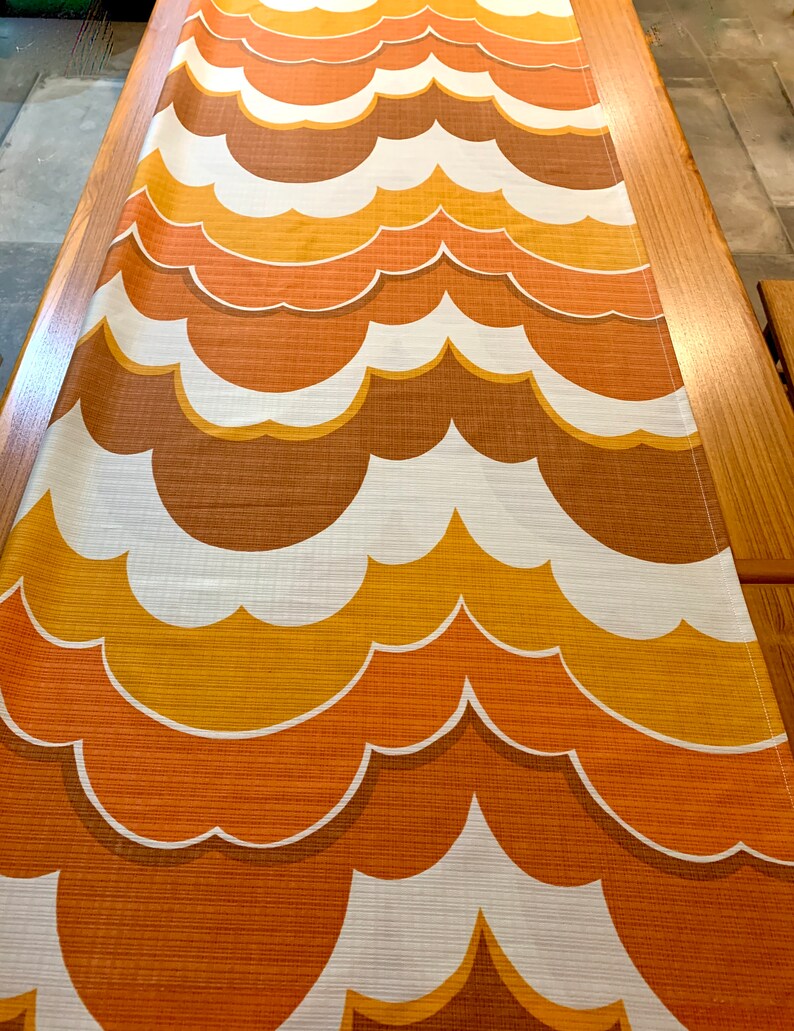 Op de afbeelding: Een tafelloper met een herhalend patroon van wolkvormige vormen in bruin-, oranje- en wittinten. De loper is gemaakt van een textuurstof en wordt gepresenteerd op een houten tafel. Het ontwerp roept een vintage esthetiek op.