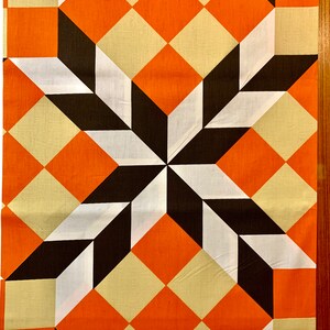 K&ouml;nnte beinhalten: Ein geometrischer Stoff mit einem sich wiederholenden Muster aus orangefarbenen, wei&szlig;en und schwarzen Quadraten. Die Quadrate sind diagonal angeordnet und erzeugen so eine Tiefenwirkung und Bewegung.