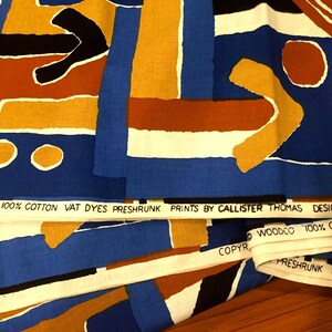 May include: Close-up of fabric with a geometric pattern in shades of blue, orange, yellow, white, and black. The fabric has a label that reads "WOODCO 100% COTTON VAT DYES PRESHRUNK PRINTS BY CALLISTER THOMAS DESIGN "HONGO WOODCO 100% COTTON VAT DYE COPYR FRESHRUNK PRINTS BY CA".