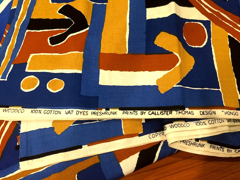 May include: Close-up of fabric with a geometric pattern in shades of blue, orange, yellow, white, and black. The fabric has a label that reads "WOODCO 100% COTTON VAT DYES PRESHRUNK PRINTS BY CALLISTER THOMAS DESIGN "HONGO WOODCO 100% COTTON VAT DYE COPYR FRESHRUNK PRINTS BY CA".