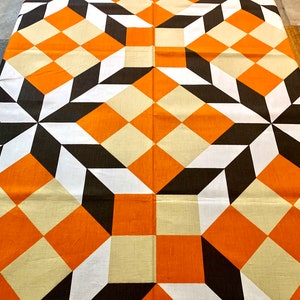 K&ouml;nnte beinhalten: Ein Stoff mit geometrischem Muster in Orange, Wei&szlig;, Schwarz und Beige mit Quadraten und Rauten. Der Stoff hat ein sich wiederholendes Muster aus Quadraten und Rauten.